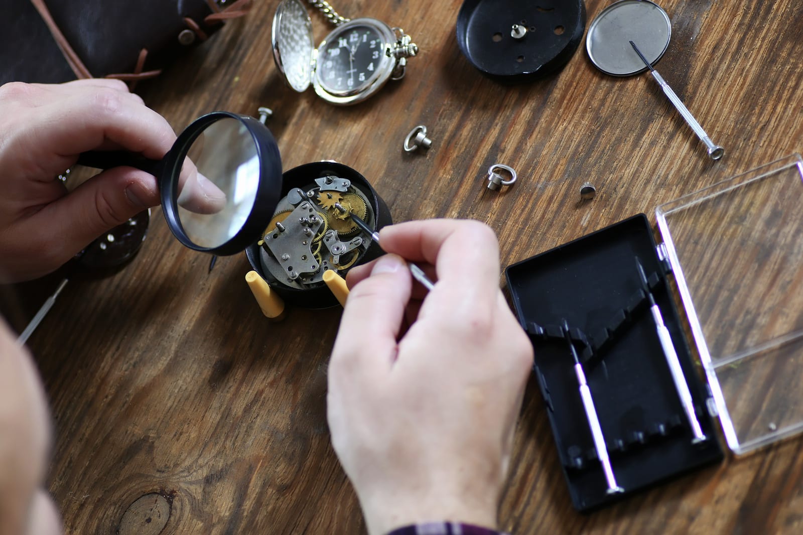Mani di un orologiaio che osservano con una lente un movimento a tasca aperto su un banco di legno